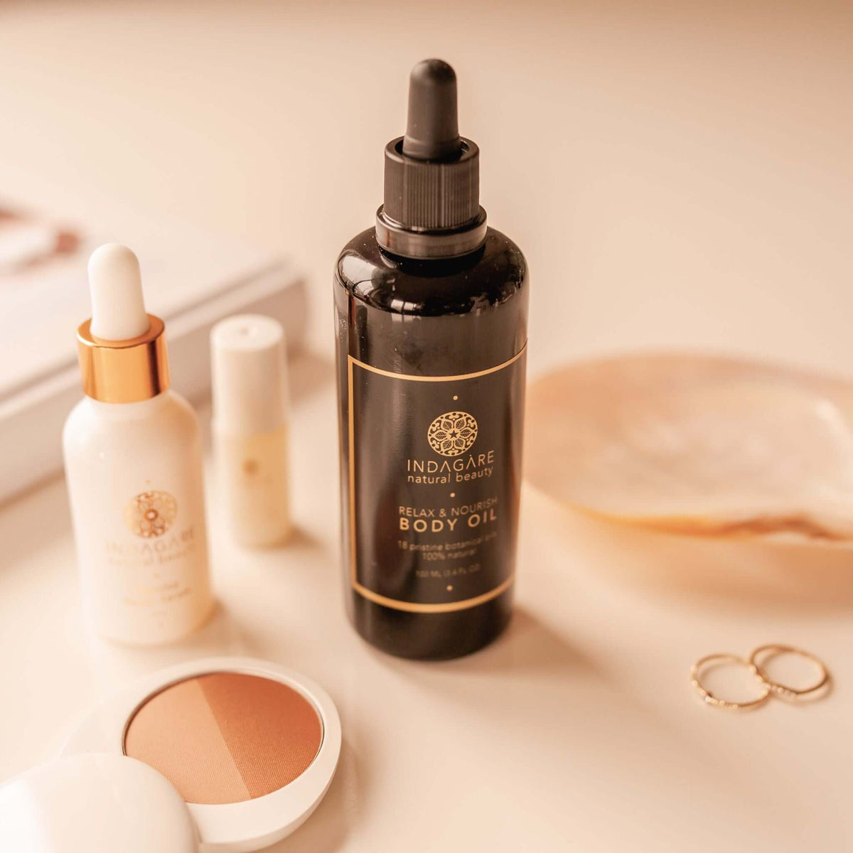Indagare's Relax and Nourish Organic Body Oil displayed on a bathroom vanity with hair oil and makeup products